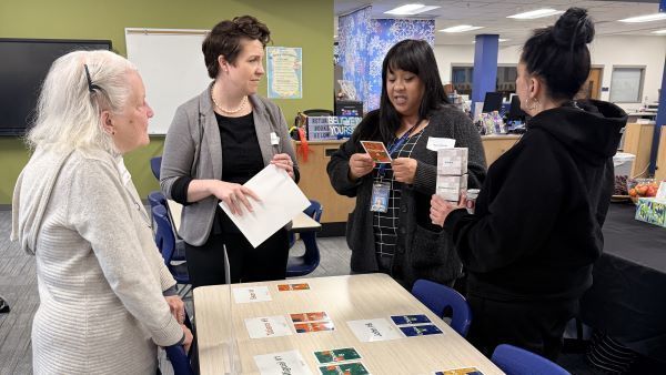 Participants collaborating during a hands-on simulation. The group is shown reviewing "Choice" cards and organizing materials at a designated station in a modern classroom or office space.