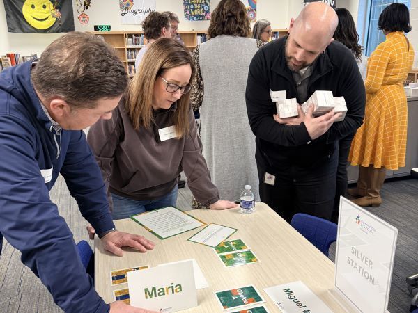 Two women and a man lean over a table filled with activity cards and name signs like "Maria." One man holds a stack of white boxes while the group examines "Choice A" cards during a simulation. In the background, other participants stand near a bookshelf in a brightly lit room.
