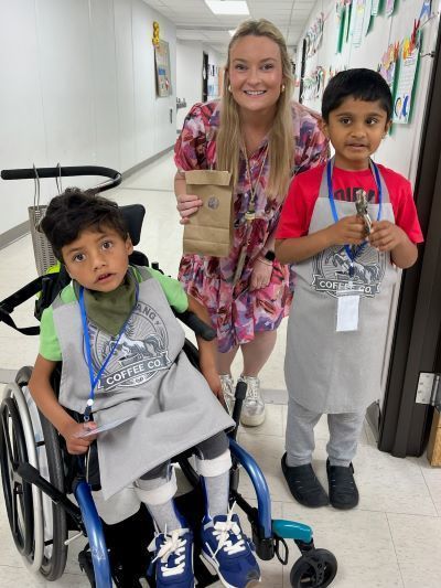 Two students in "Wild Mustang Coffee Co." aprons and their teacher posing in a school hallway.