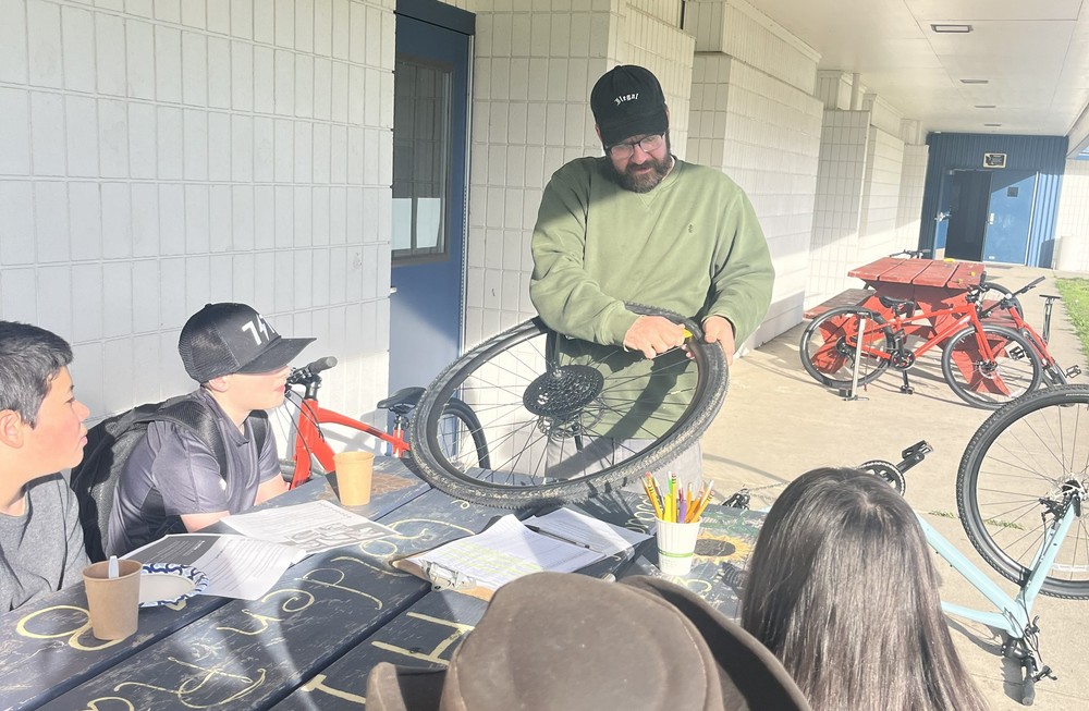 ReCyclery Instructor holding a tire and showing how to make a repair, students sitting at table, outdoors.