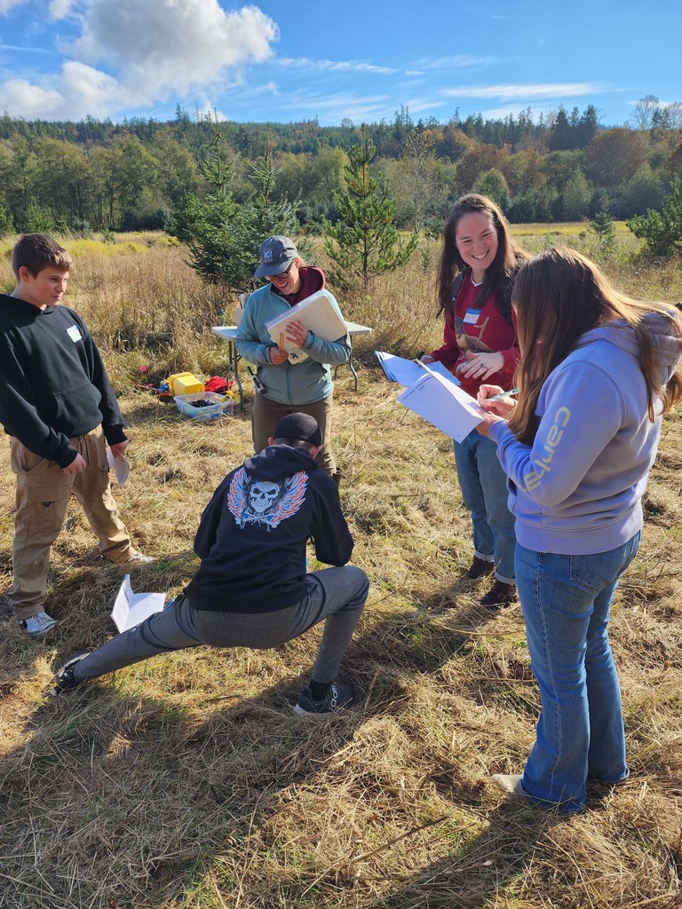 Two teachers and three students standing outdoors under blue sky, looking at notebooks, and studying nature.