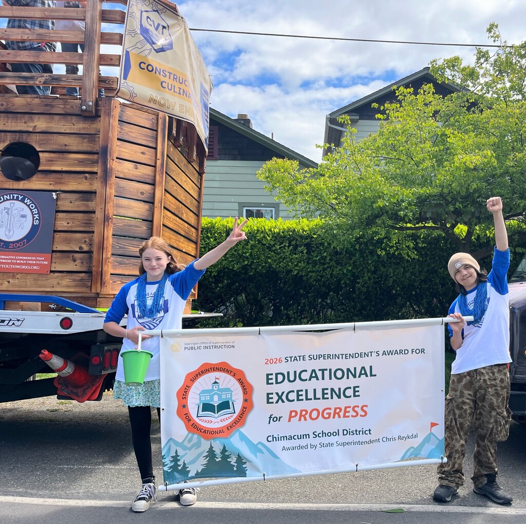 Two students celebrating with fists in the air while holding an Educational Excellence Banner for Progress award banner in the Rhody Parade.