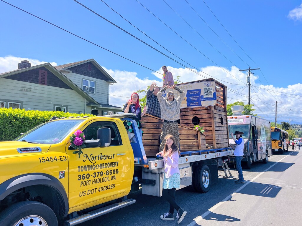 North West Tow truck pulling the flatbed trailer with the student built ship and six kids waving, one making a heart shape with his hand.