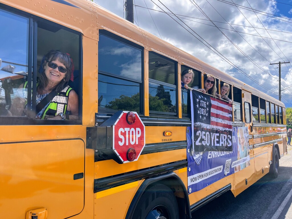 Chimacum School Bus Driver Judy smiling through the bus window, her daughter and 2 girls smiling and holding an American flag out the window.