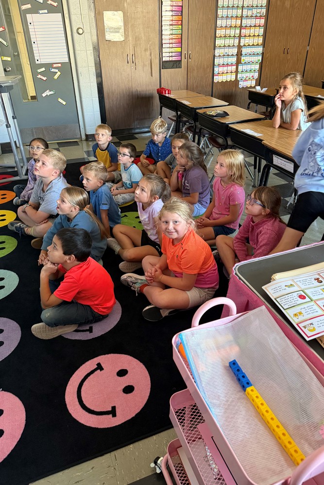 Numerous second grade students are sitting on a large black rug with large colored smiley faces