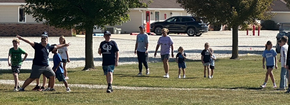Students and their families are walking outside in front of tall , green trees