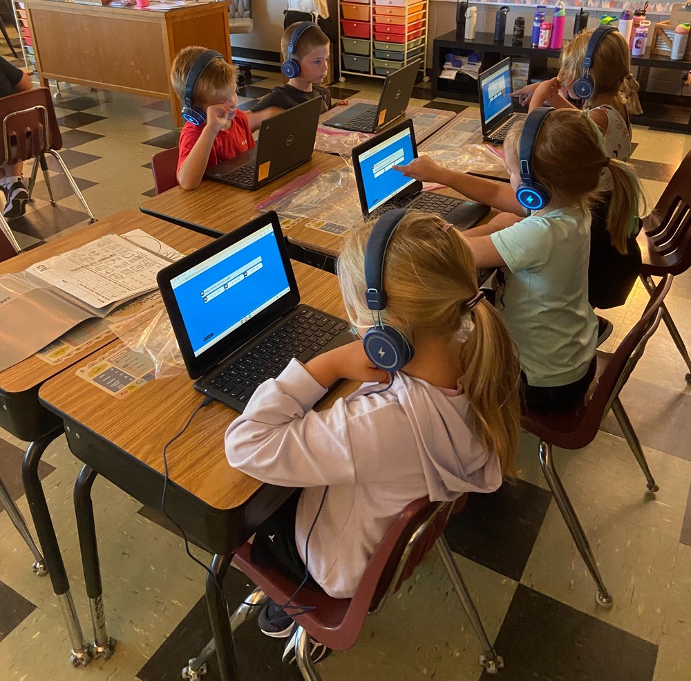 Five first grade students are sitting at desks with headphones and chromebooks