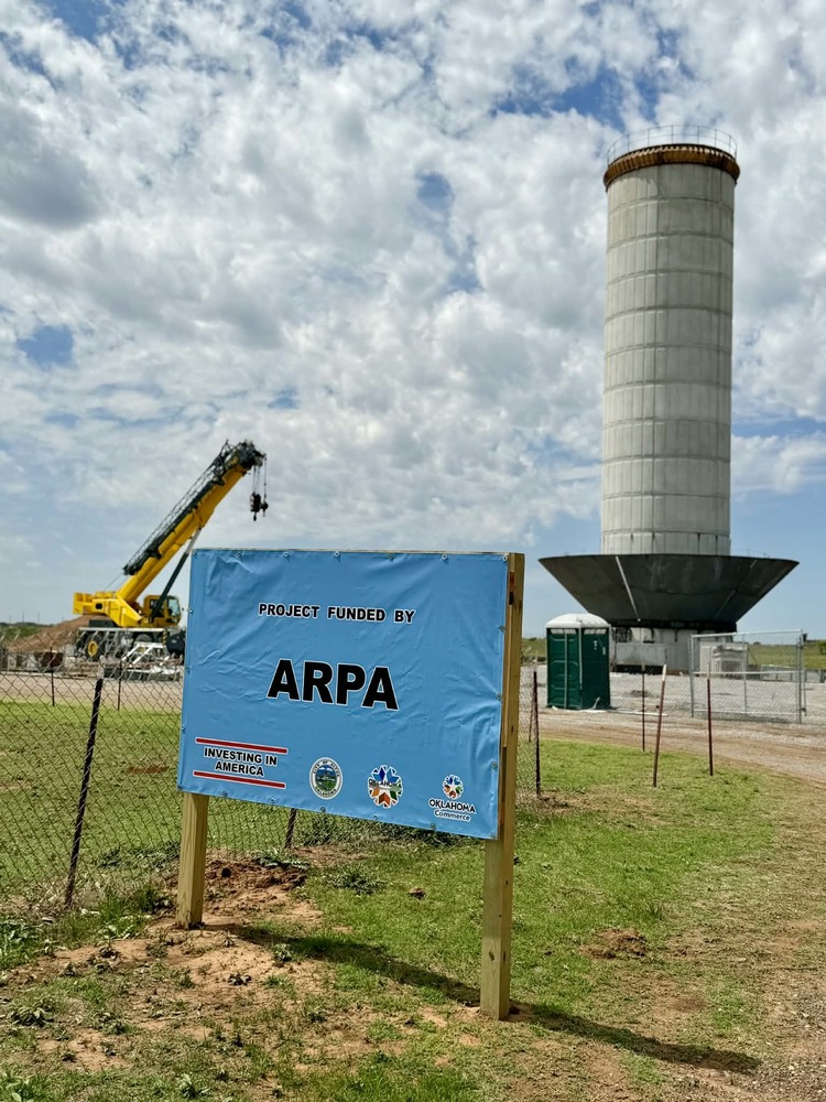 A tall white water tower under construction rises above a fenced work site, with a crane and equipment in the background and a blue sign in the foreground indicating the project is funded by ARPA.