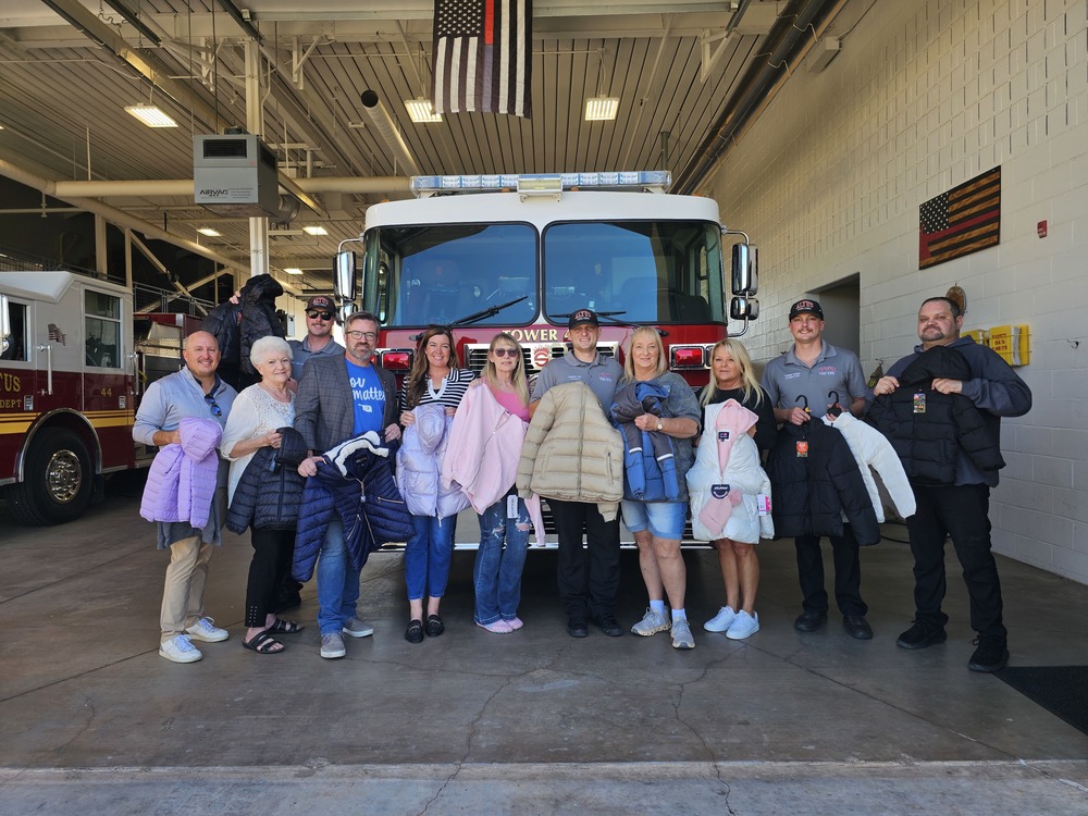 Group of Altus Fire and EMS staff and community members pose in a fire station bay in front of a fire engine, holding winter coats for the Coats for Kids drive.