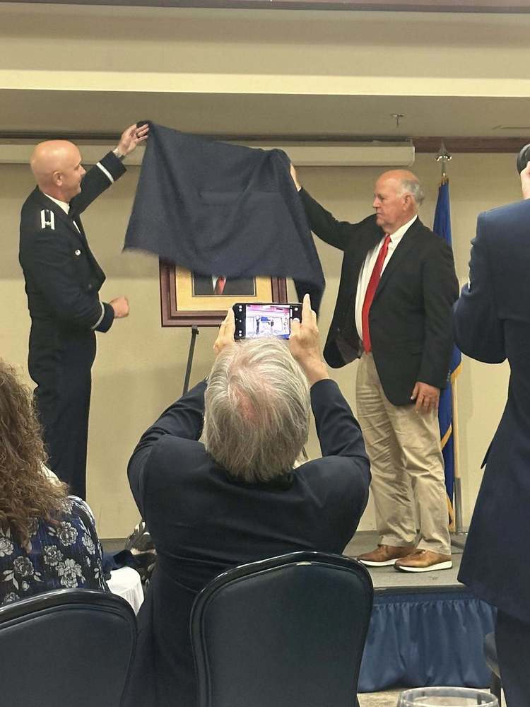 City Manager Gary Jones stands on stage beside an Air Force officer as they unveil a framed portrait, while an audience member captures the moment on a phone