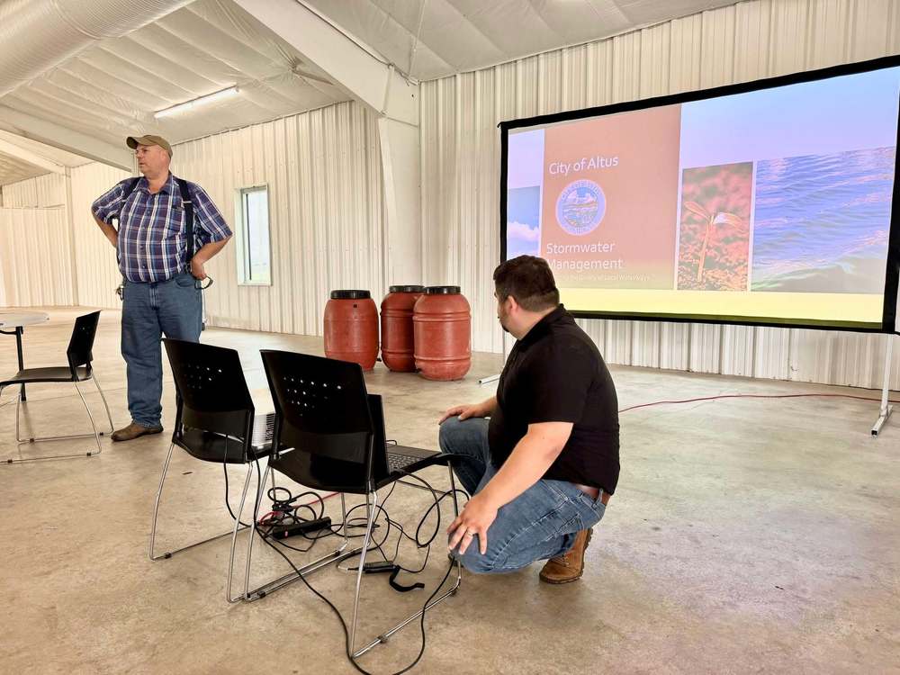 Two men setting up for a City of Altus stormwater management presentation inside a white metal building. A projector screen displays the City of Altus logo and presentation slide.