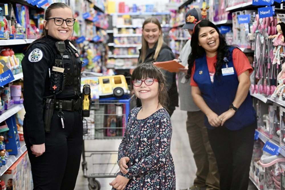 A police officer and Walmart employees smile while shopping with a young child in a store aisle during the Cops and Kids Christmas event.