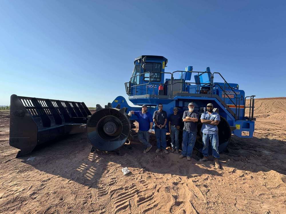 Kyle Pipho and members of the Altus Landfill Department stand in front of the new blue Aljon 500 compactor on a dirt worksite under a clear blue sky.