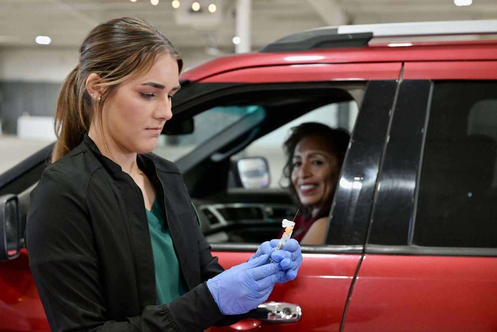 A healthcare worker wearing gloves prepares a flu shot while standing beside a red SUV during a drive-thru vaccination event. A person in the driver’s seat smiles from the car window.