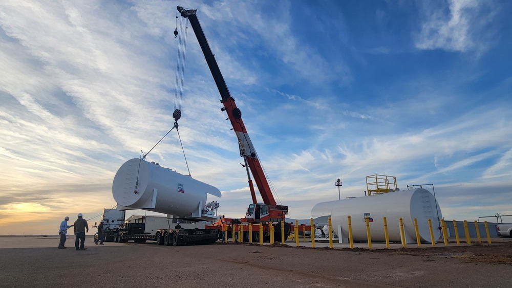 Crane lifting a large white fuel storage tank onto a trailer on the airport tarmac at sunset, with workers nearby and another fuel tank installed on a concrete pad in the background.