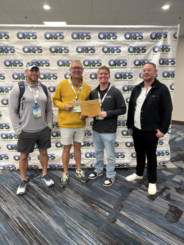 Four Altus Parks and Recreation staff members stand together in front of an Oklahoma Recreation and Parks Society step and repeat backdrop. From left to right are Todd Kirouac, Michael Shive, Tyler Gray holding the award plaque, and Eric Warren. All four are smiling and dressed in conference attire.