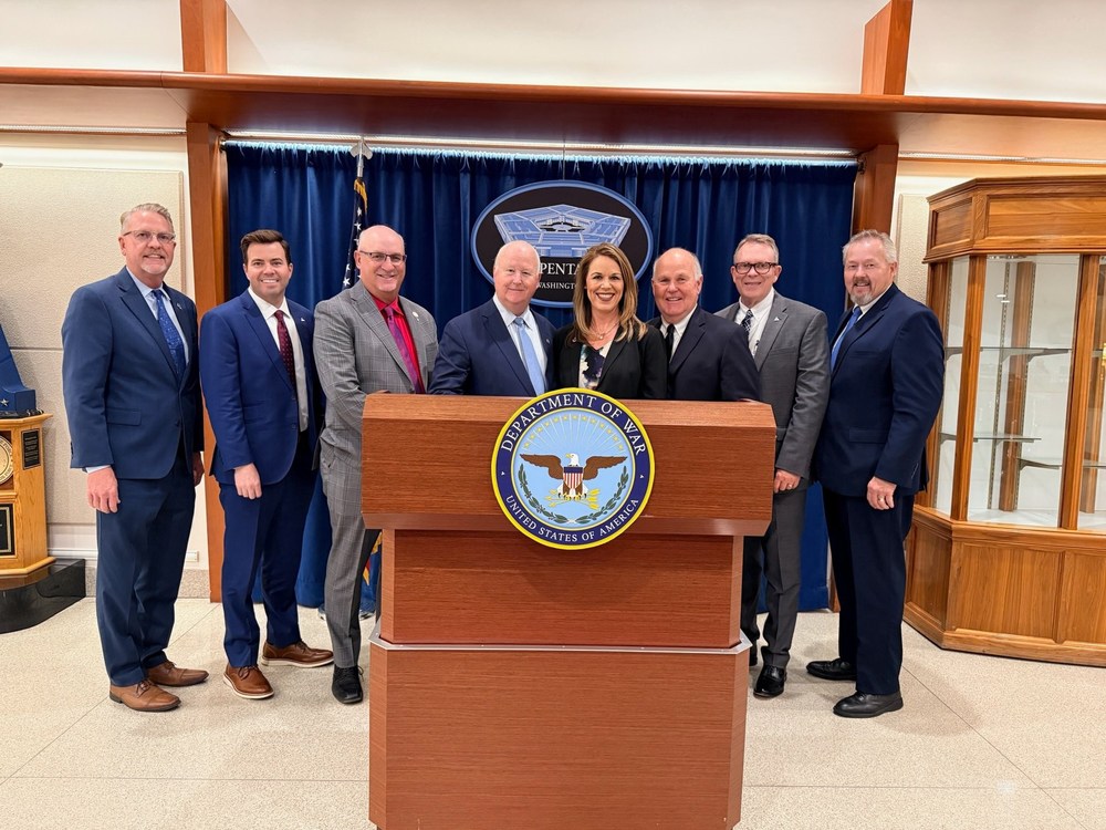 A group of community leaders and officials stand behind a podium with a Department of War seal, posing for a photo in a formal room.