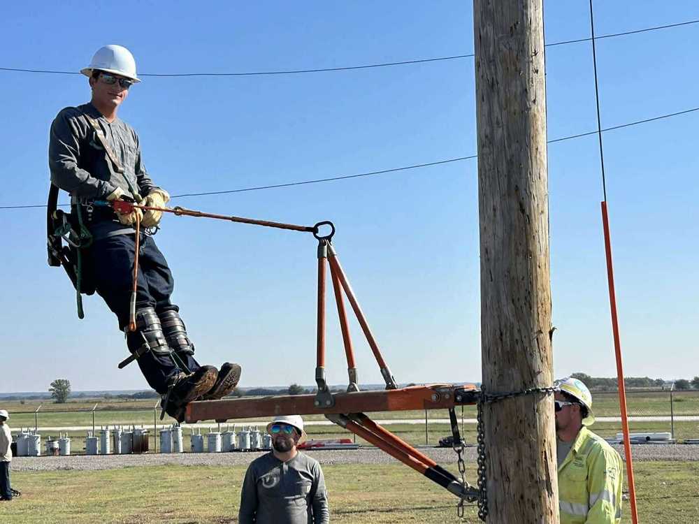 A worker wearing safety gear practices climbing and balancing on a utility pole during a lineman training exercise, with two other workers observing nearby.