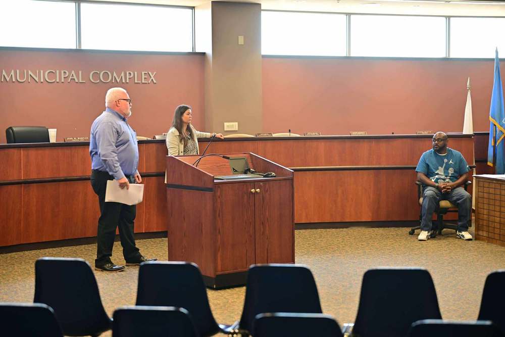 hree people inside a municipal courtroom during a training session. Two individuals stand near the podium, one holding papers, while another is seated to the side. The room features a wood-paneled dais, microphones, flags, and rows of empty chairs in the foreground.