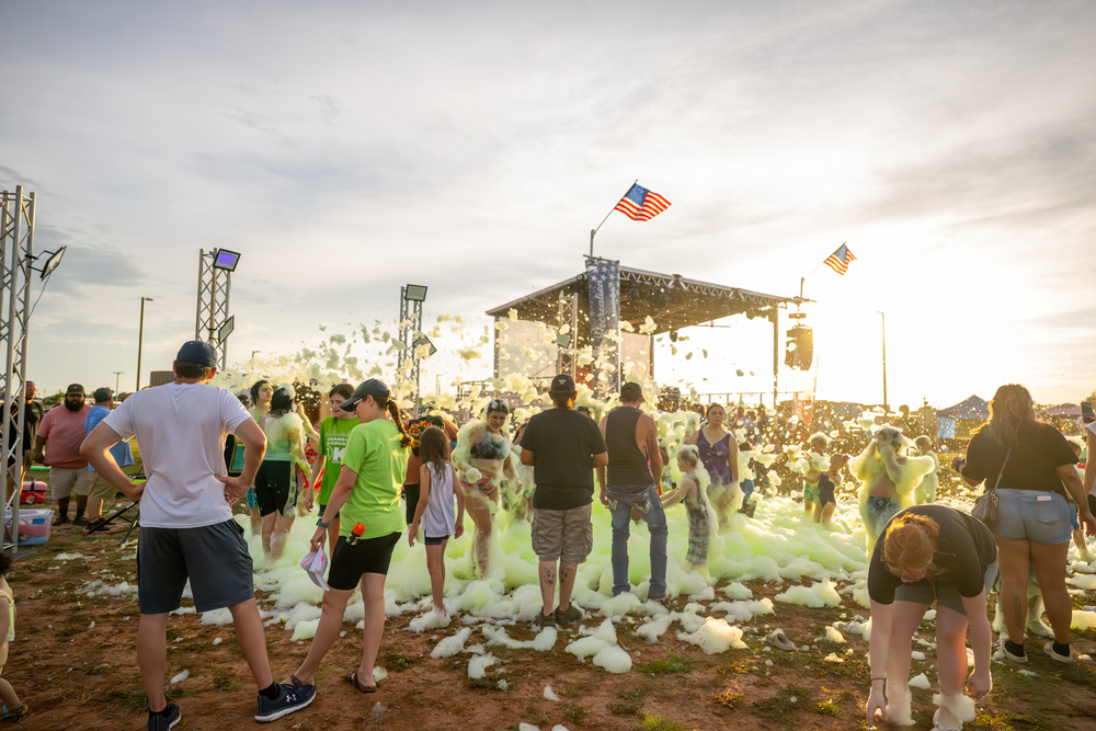Festival-goers of all ages enjoy a foam party in front of a concert stage at sunset, with American flags flying above the stage at the 2025 Altus Spirit Festival.