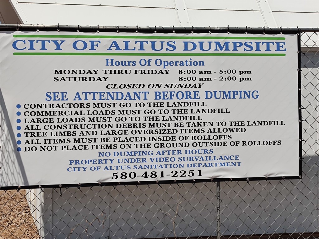 City of Altus Dumpsite sign showing hours of operation and rules. Open Monday to Friday 8 a.m. to 5 p.m., Saturday 8 a.m. to 2 p.m., closed Sunday. Sign instructs contractors, commercial loads, and large loads to go to the landfill. Tree limbs and oversized items allowed but must be placed inside roll-off containers. No dumping after hours. Property under video surveillance. City of Altus Sanitation Department phone number 580-481-2251