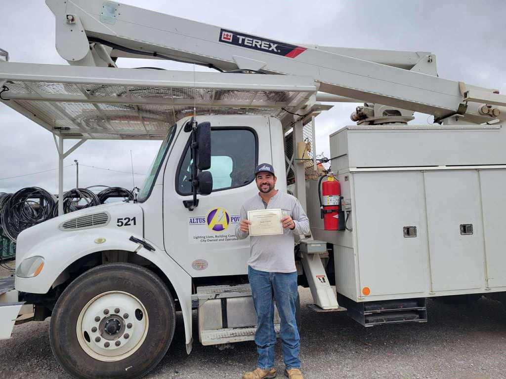 Jesse Harris, Altus Power employee, stands in front of a white utility bucket truck holding his Journeyman Lineman certificate and smiling proudly.