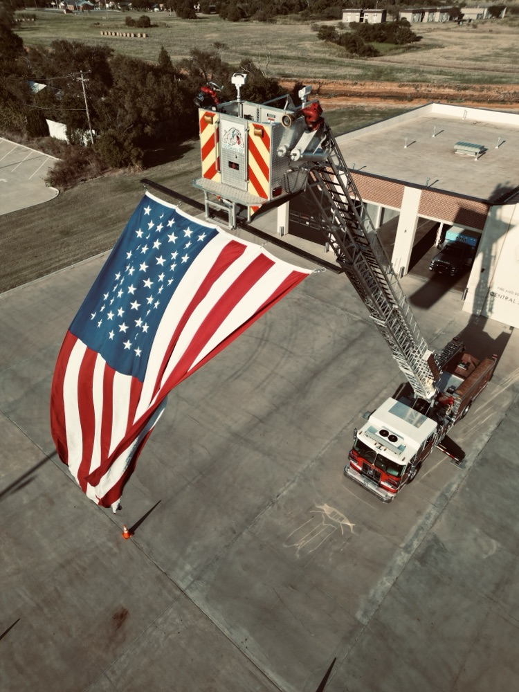 Large American flag hanging from the extended ladder of an Altus Fire Department truck outside the fire station, displayed in honor and remembrance of September 11.