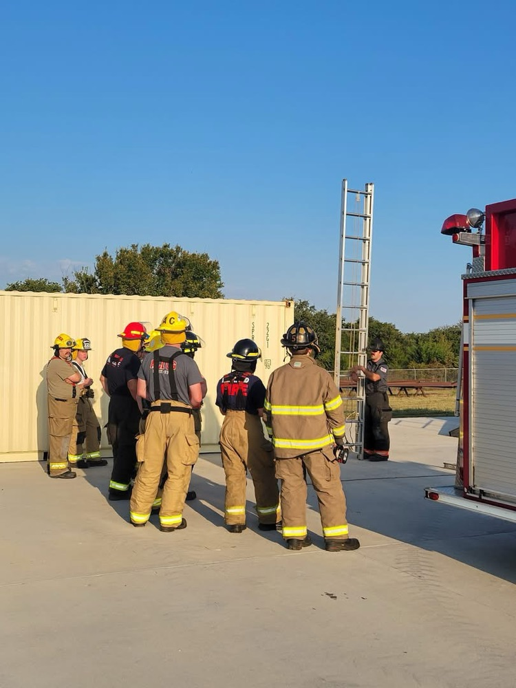 Group of firefighters in full gear stand in a circle listening to an instructor demonstrating ladder techniques next to a fire engine and training containers.