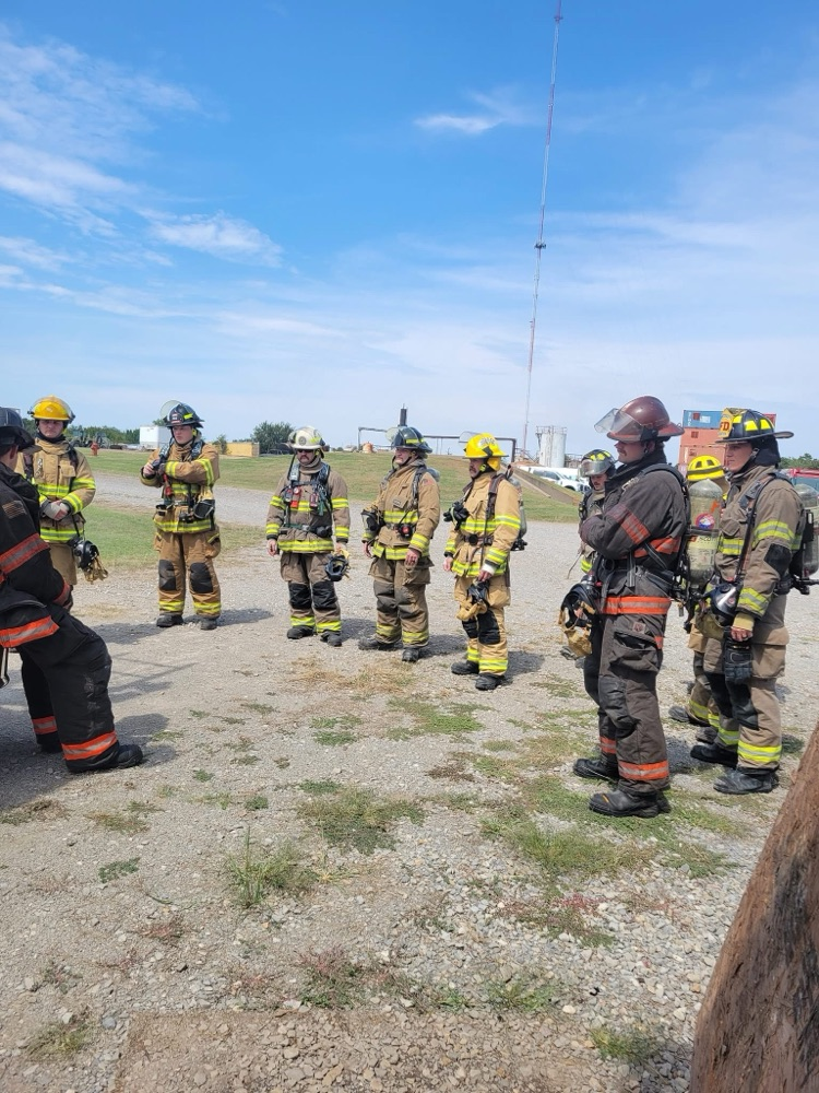 Group of firefighters in full gear stand in a circle listening to an instructor demonstrating ladder techniques next to a fire engine and training containers.