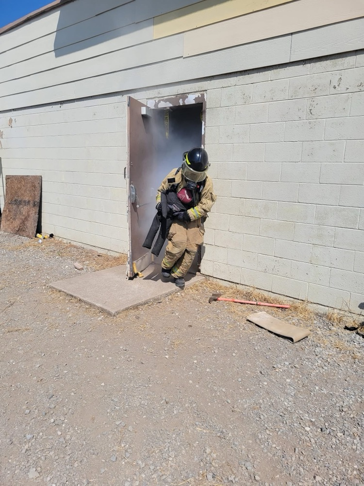 Firefighter in full gear and breathing mask emerging from a smoke-filled doorway carrying a training dummy, with tools on the ground nearby.