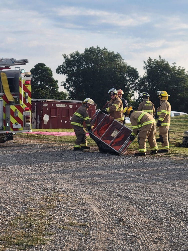 Group of firefighters in full turnout gear lifting a large red training container next to a fire engine during outdoor training.