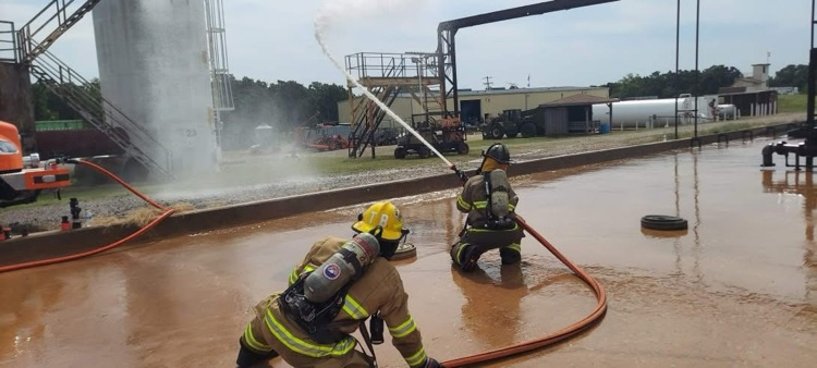 Two firefighters in yellow helmets kneeling and spraying a powerful stream of water from a fire hose toward an outdoor training structure with tanks and pipes.