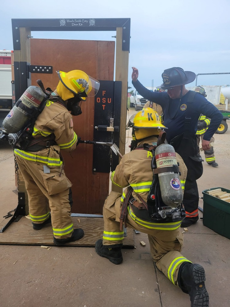 Firefighters in turnout gear practicing forcible entry on a wooden door prop under the supervision of an instructor in a black helmet.