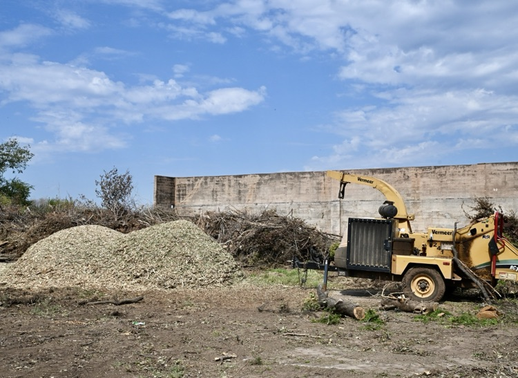 A large pile of wood mulch sits at the Blaine Drop-Off Site in Altus, with a yellow Vermeer wood chipper parked beside it under a partly cloudy sky.