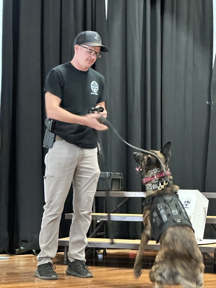 A K9 officer stands on stage holding the leash of a police dog wearing a tactical vest. The dog looks up attentively at the handler during the demonstration.