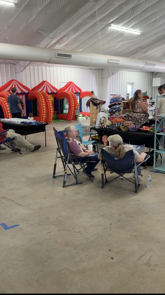 Inside a large event space, people sit in foldable chairs near vendor tables displaying handmade items. Inflatable carnival-style play structures are set up in the background.