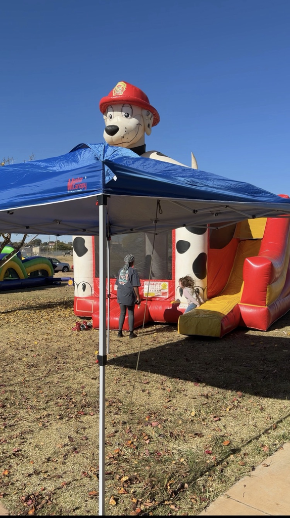 A large inflatable bounce house decorated like a Dalmatian firefighter sits on a grassy area. A blue canopy tent is set up in front, and two people stand near the entrance of the bounce house.