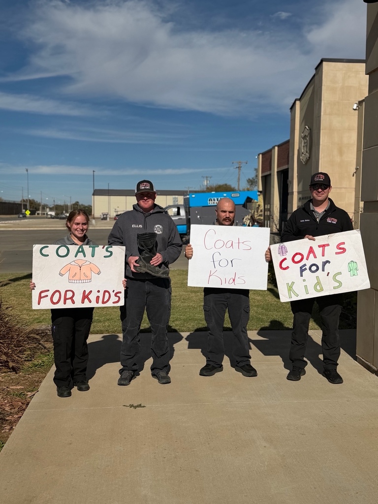 Four Altus Fire EMS personnel standing outside the fire station holding handmade signs that say āCoats for Kids.ā They are smiling and standing on the sidewalk in front of the station on a sunny day.