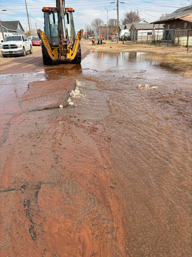 Water flowing across a roadway with city utility equipment and crews working in the background near a residential area.