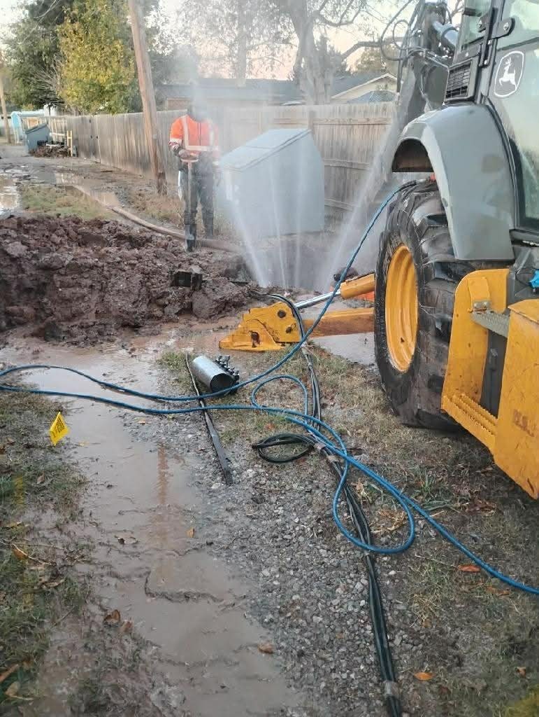 City of Altus utility crew repairing a broken water main as water sprays from the damaged line near heavy equipment.