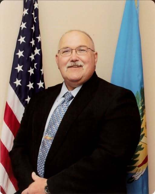 Image of Robert "Bob" McGill  wearing a suit and tie, standing in front of the american and oklahoma flag. 