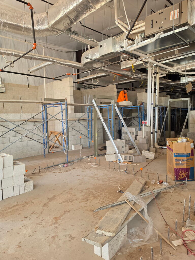 A series of 5 photos showing the interior construction progress of Fire Station No. 3 in Altus. The images feature newly built concrete block walls forming future offices, bedrooms, and work areas. Exposed HVAC ductwork, fire sprinkler piping, electrical systems, and plumbing are visible overhead. Construction materials, scaffolding, ladders, and cinder blocks are positioned throughout the building. One photo shows a construction worker laying concrete blocks while working from scaffolding. Another shows a nearly completed room with finished block walls and a concrete floor. The final image provides a wide view of the station's interior, where room layouts are clearly visible as crews prepare for the next phase of construction, including exterior brick installation. Substantial completion of the project is expected in September 2026.
