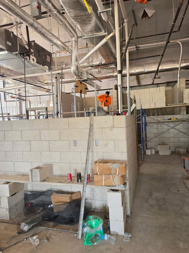 A series of 5 photos showing the interior construction progress of Fire Station No. 3 in Altus. The images feature newly built concrete block walls forming future offices, bedrooms, and work areas. Exposed HVAC ductwork, fire sprinkler piping, electrical systems, and plumbing are visible overhead. Construction materials, scaffolding, ladders, and cinder blocks are positioned throughout the building. One photo shows a construction worker laying concrete blocks while working from scaffolding. Another shows a nearly completed room with finished block walls and a concrete floor. The final image provides a wide view of the station's interior, where room layouts are clearly visible as crews prepare for the next phase of construction, including exterior brick installation. Substantial completion of the project is expected in September 2026.