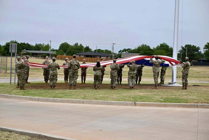 Uniformed service members stand in a circle holding a large American flag near the Altus Reservoir Roundabout flagpole.