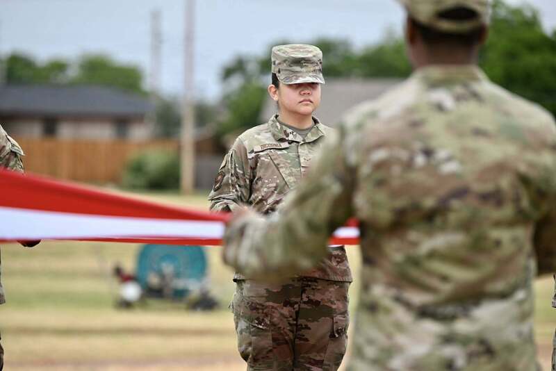 A uniformed service member stands at attention while others hold a large American flag during the Ceremonial Raising of the Colors.