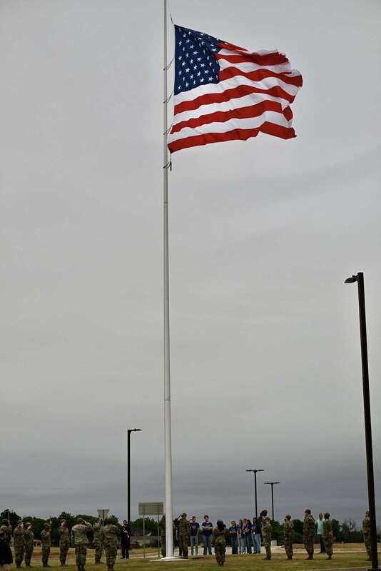 A large American flag flies from an 80-foot flagpole at the Altus Reservoir Roundabout while service members and community members stand below.