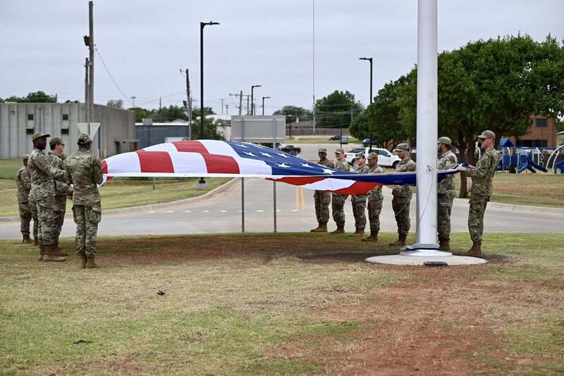 Uniformed service members hold a large American flag stretched out near the base of the flagpole before it is raised.