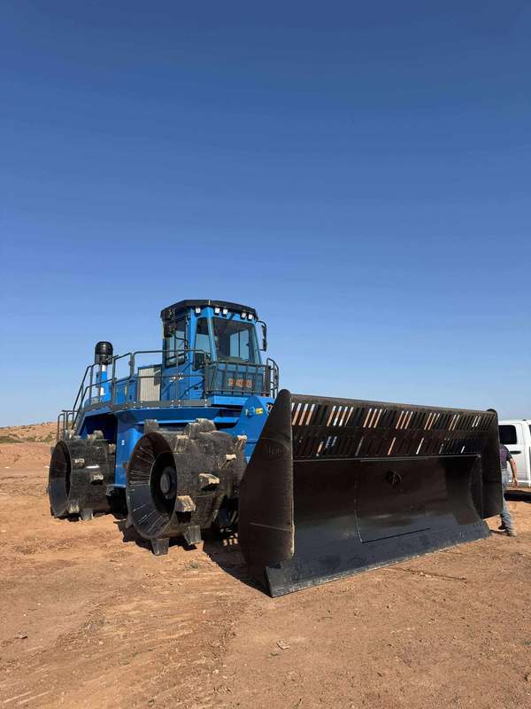 Front view of the blue Aljon 500 compactor on a dirt lot under a clear sky, showing its wide blade attachment and heavy-duty wheels.