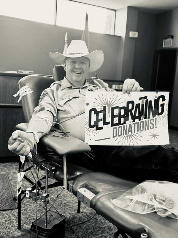 Man wearing a cowboy hat smiles while donating blood at the City of Altus Blood Drive, holding a sign that reads “Celebrating Donations!”