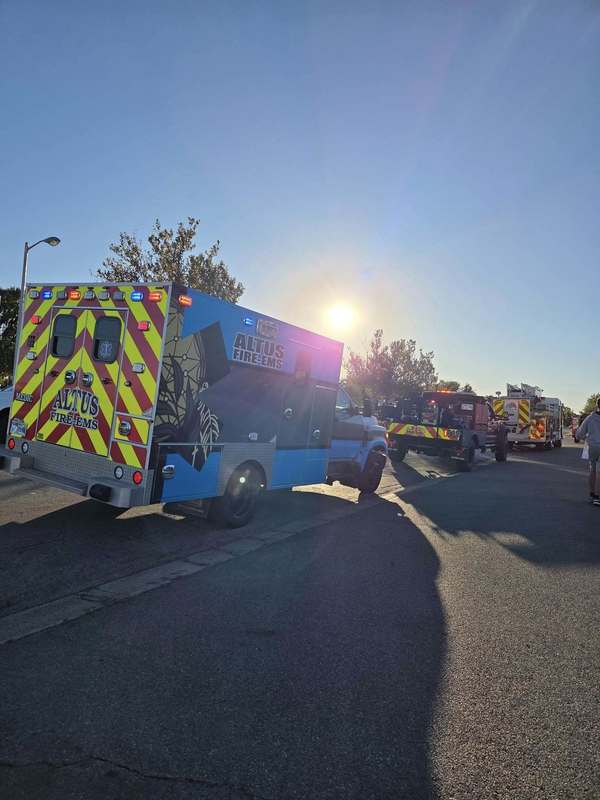 An Altus EMS ambulance and fire truck parked along a sunny street with community members gathering nearby for a birthday parade.